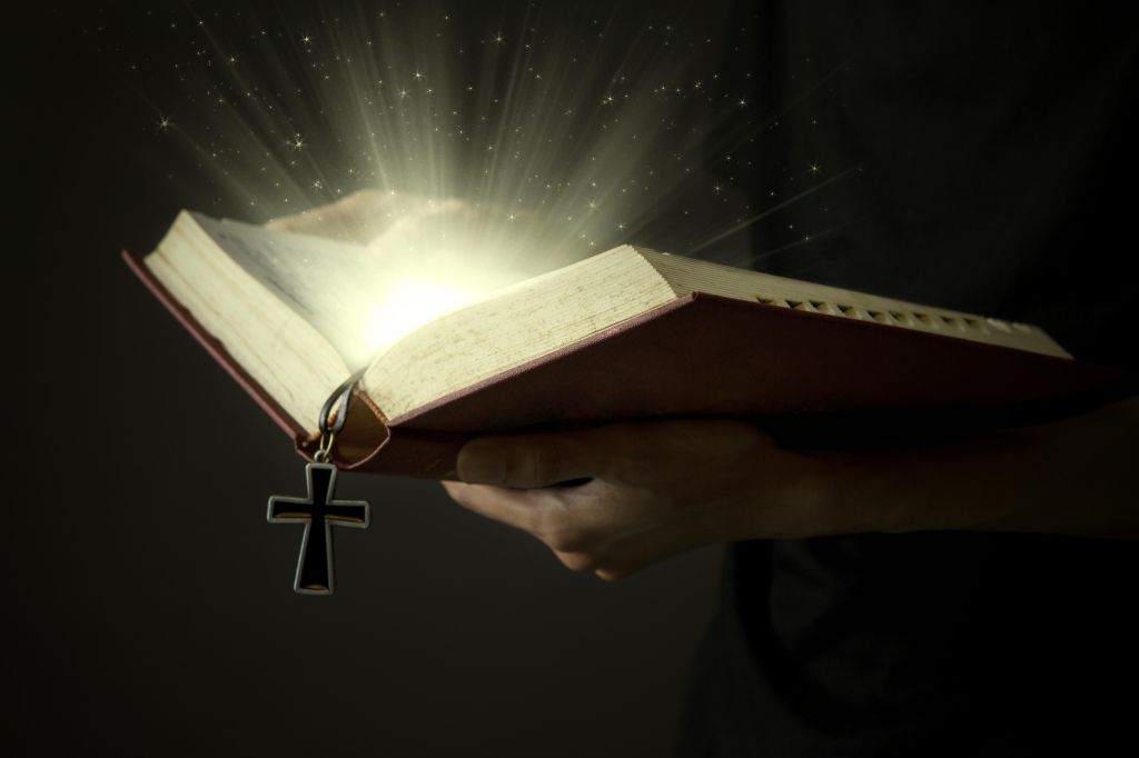 Hands of man holding holy bible and wooden rosary with magical rays