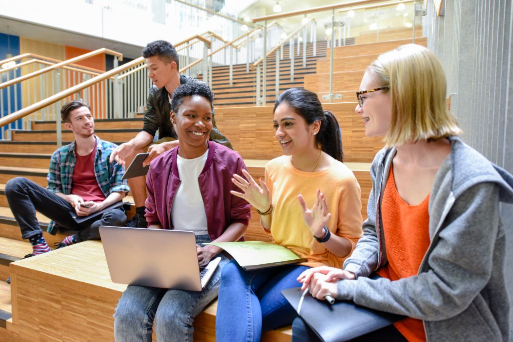 Cheerful female college friends sitting on steps with laptop and folders