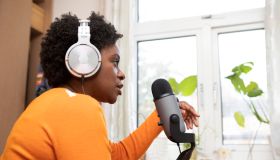 Woman at a table by a window recording a podcast home in the bedroom - with headphones and a microphone