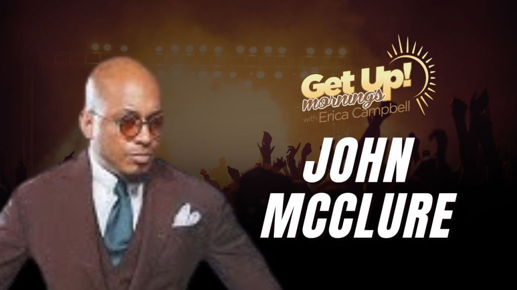 A man in a suit and glasses standing in front of a "Get Up! Mornings with Erica Campbell" backdrop with the text "JOHN MCCLURE".