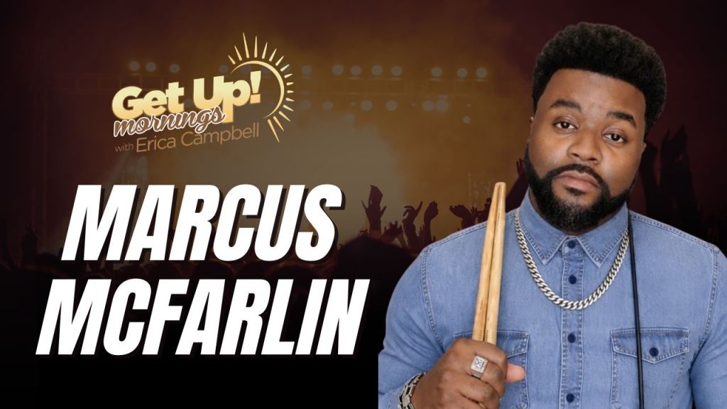 A man with a beard wearing a denim jacket and holding drumsticks stands in front of a "Get Up! Mornings with Erica Campbell" backdrop.